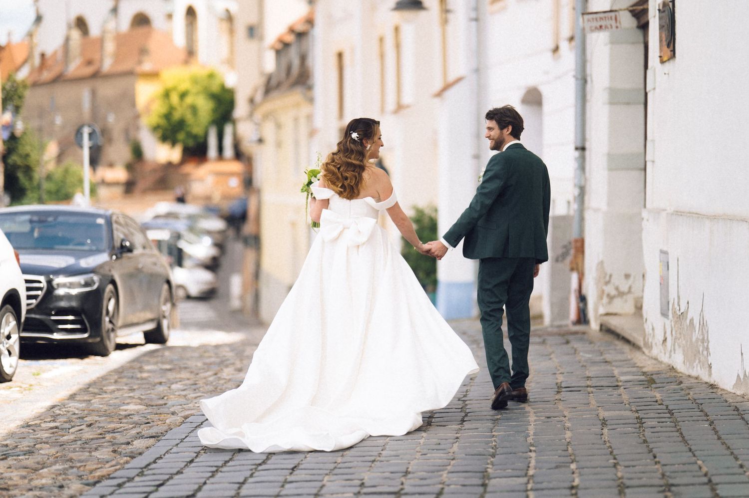 Bride in white gown and groom in dark suit walk hand-in-hand on cobblestone street in charming European town.
