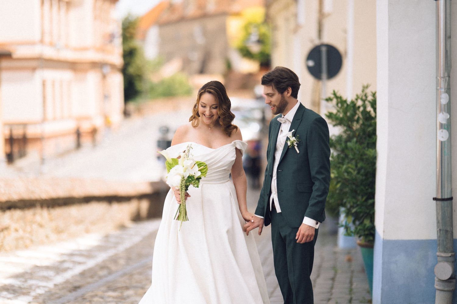 Bride in white off-shoulder gown holding bouquet walks hand-in-hand with groom in dark suit on cobblestone street.