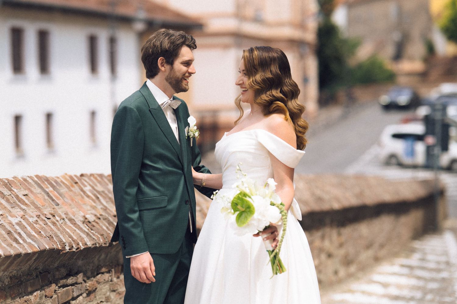 Bride and groom share a romantic moment on a cobblestone street, she holds a white bouquet in a white off-shoulder gown.