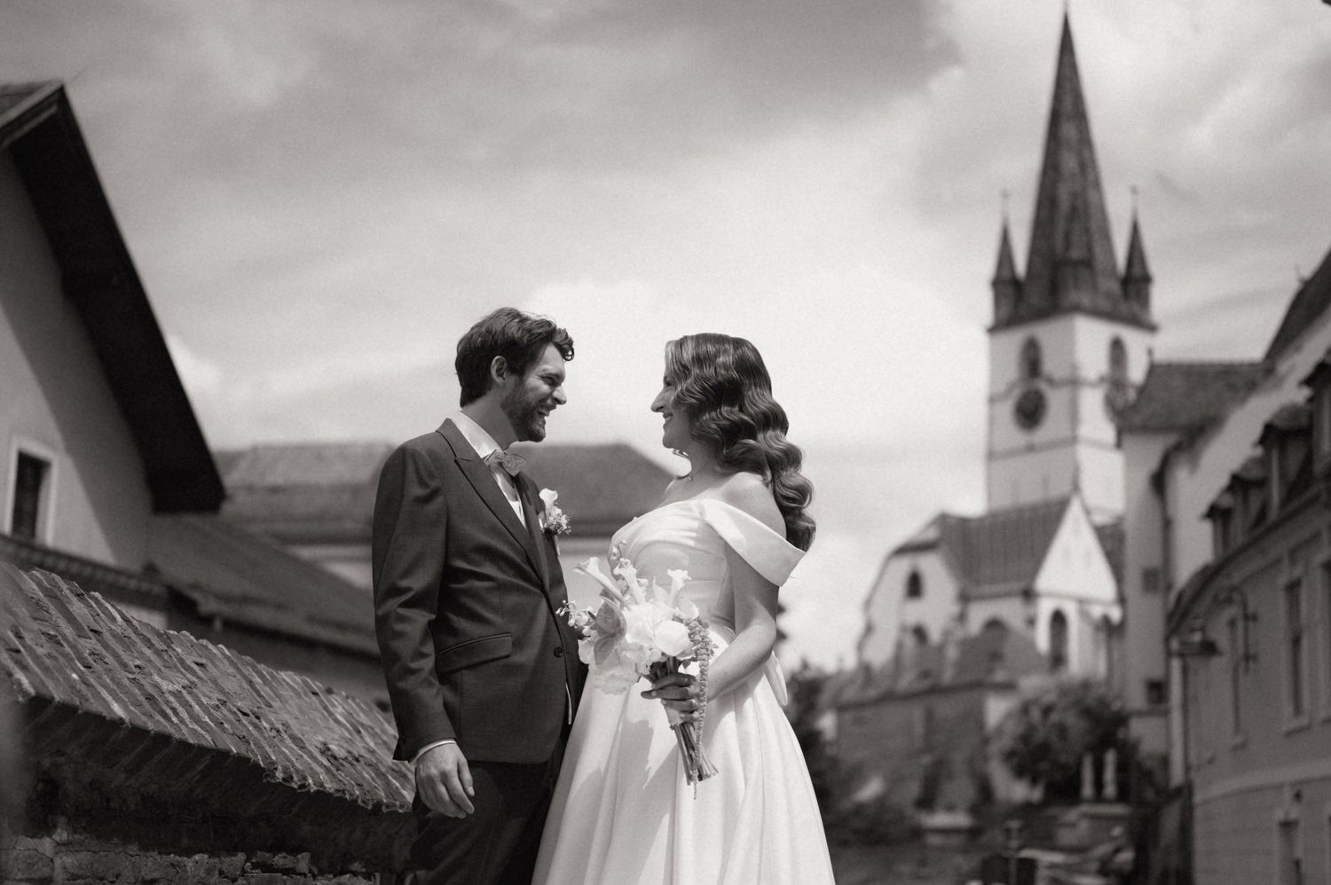 Black and white wedding photo of bride and groom standing together with a church steeple and village buildings in the background.