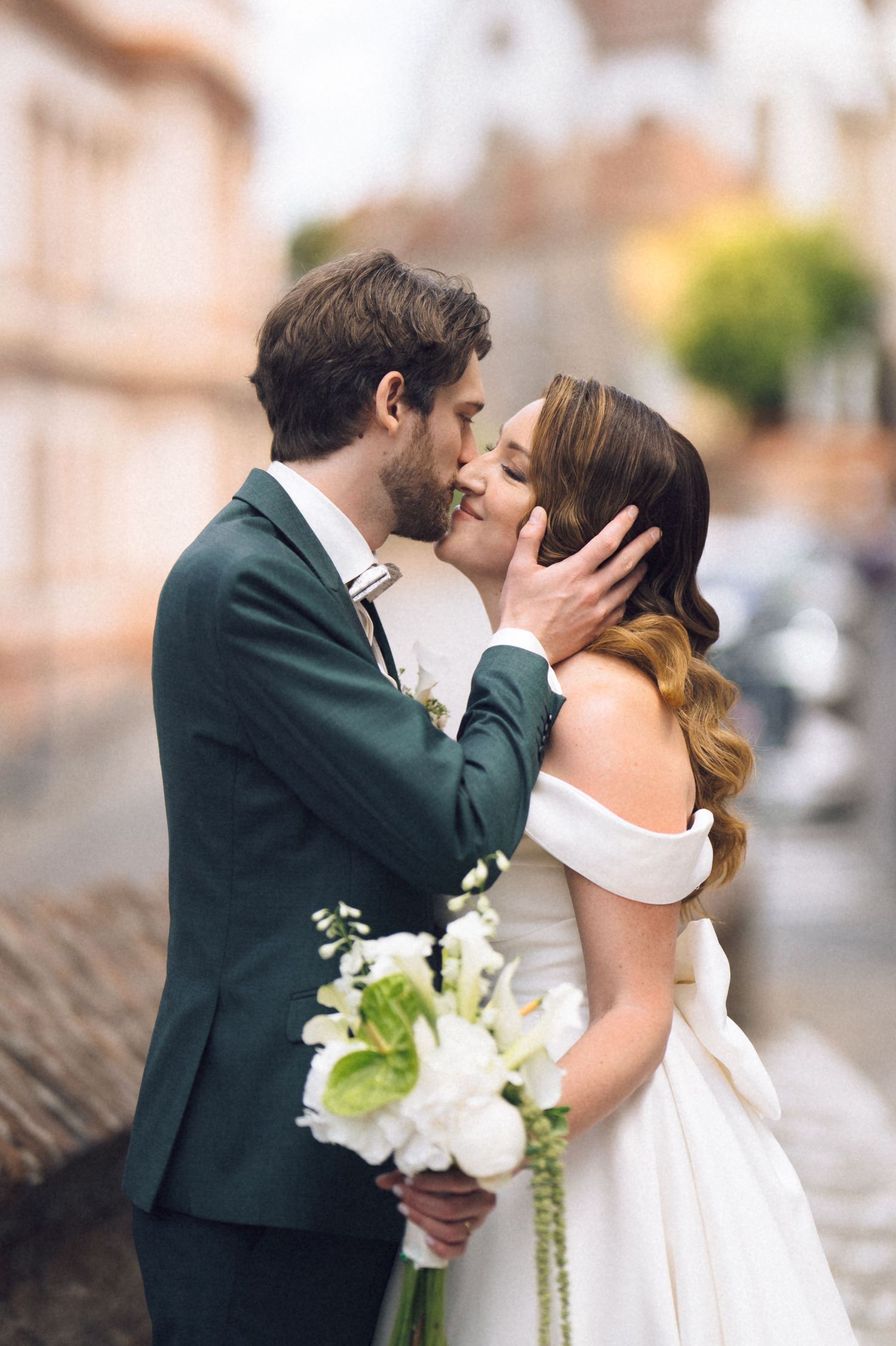 A bride and groom share a romantic kiss on a cobblestone street, bride holding white floral bouquet in off-shoulder gown.