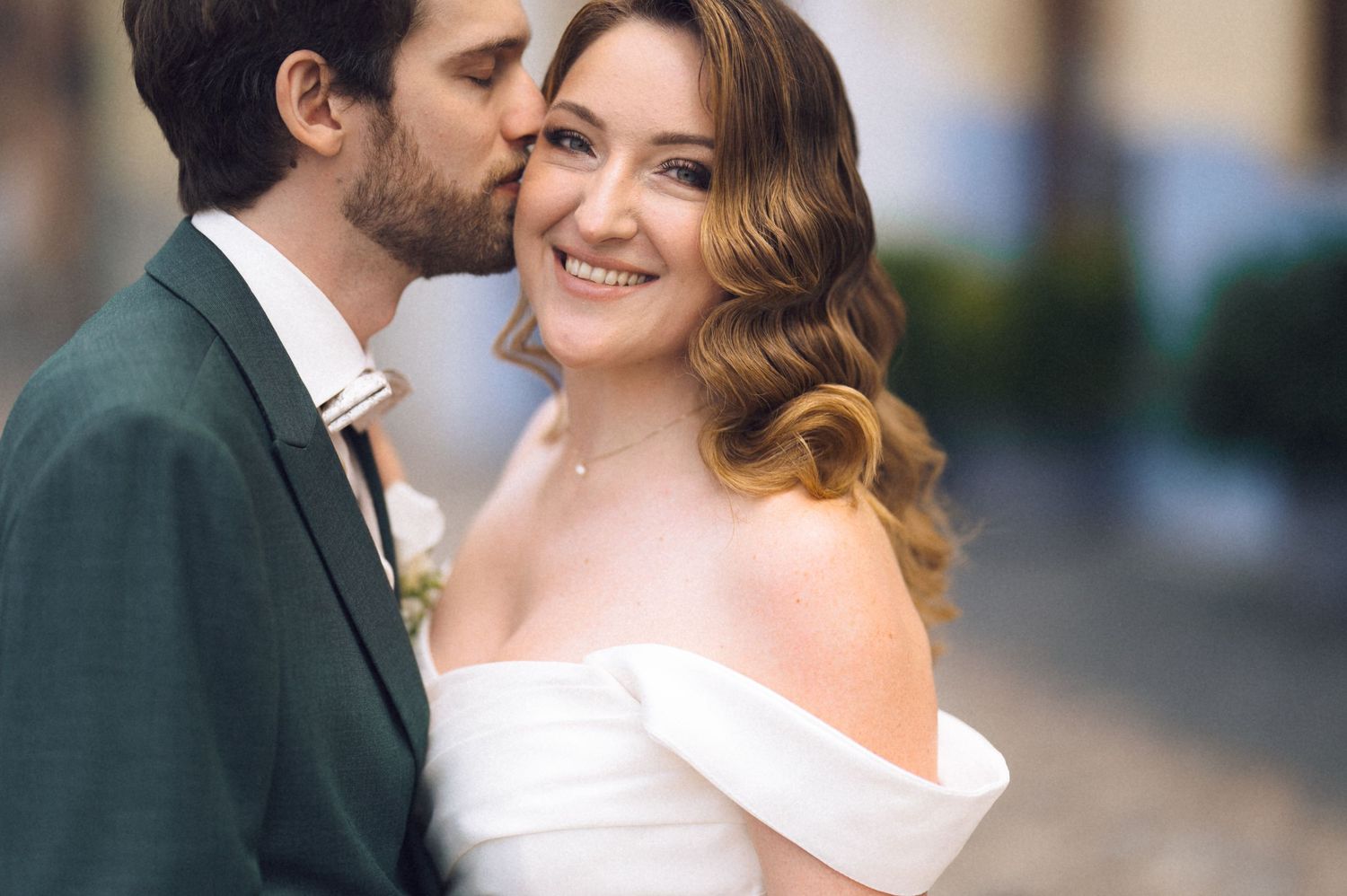 Groom in green suit kissing bride in off-shoulder white gown on cheek, smiling couple on wedding day.