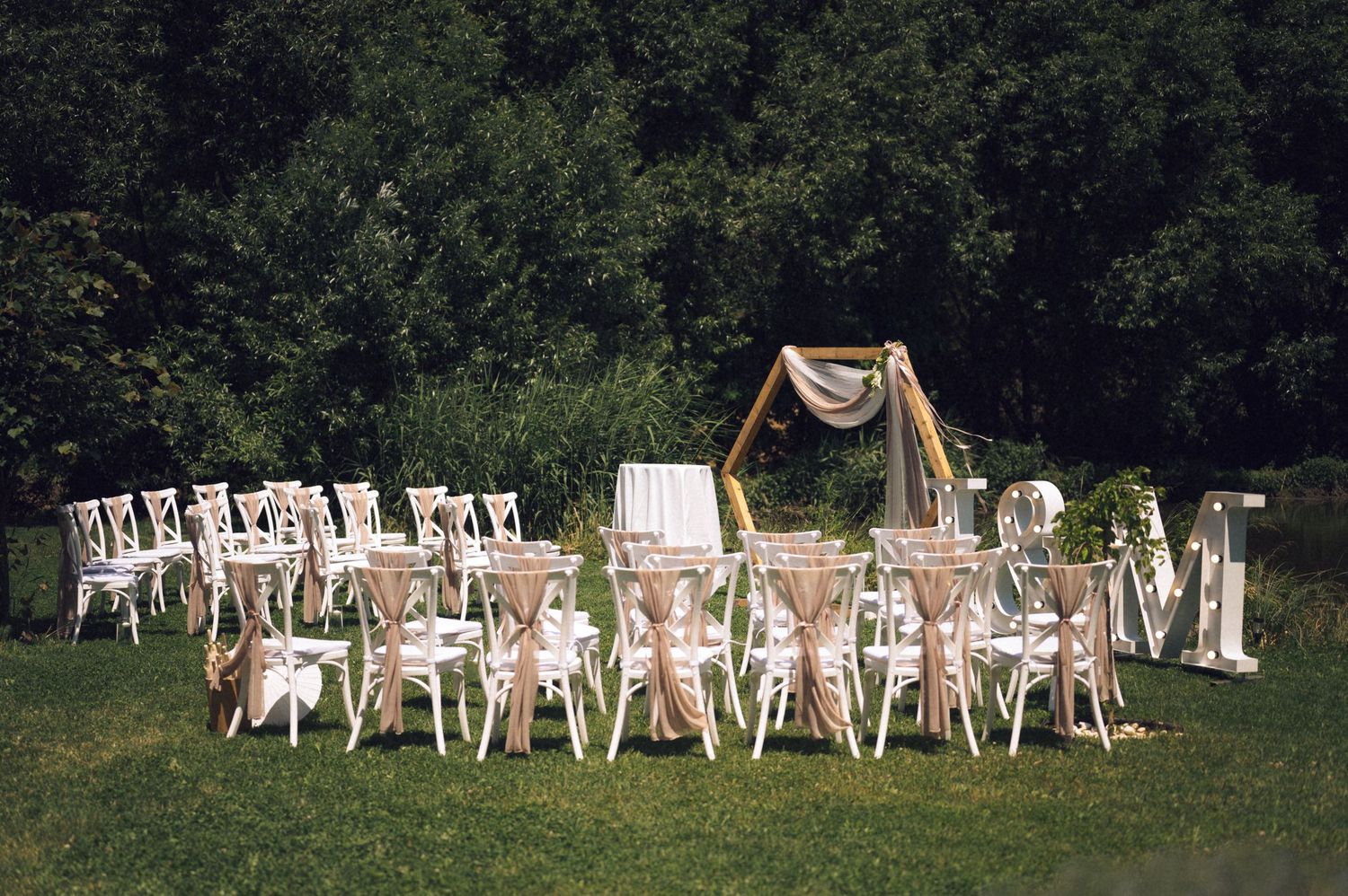 Outdoor wedding reception setup with white chairs and burlap decor on green lawn surrounded by lush trees.