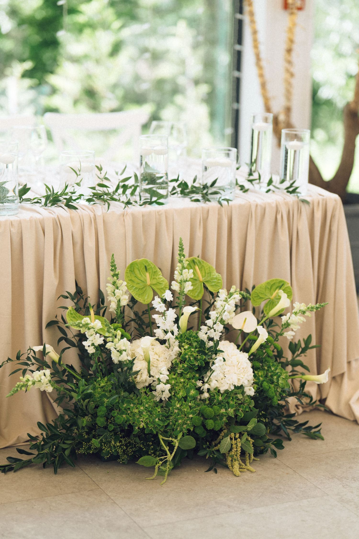 Elegant wedding reception table with beige linen, white and green floral arrangement featuring anthuriums and hydrangeas.