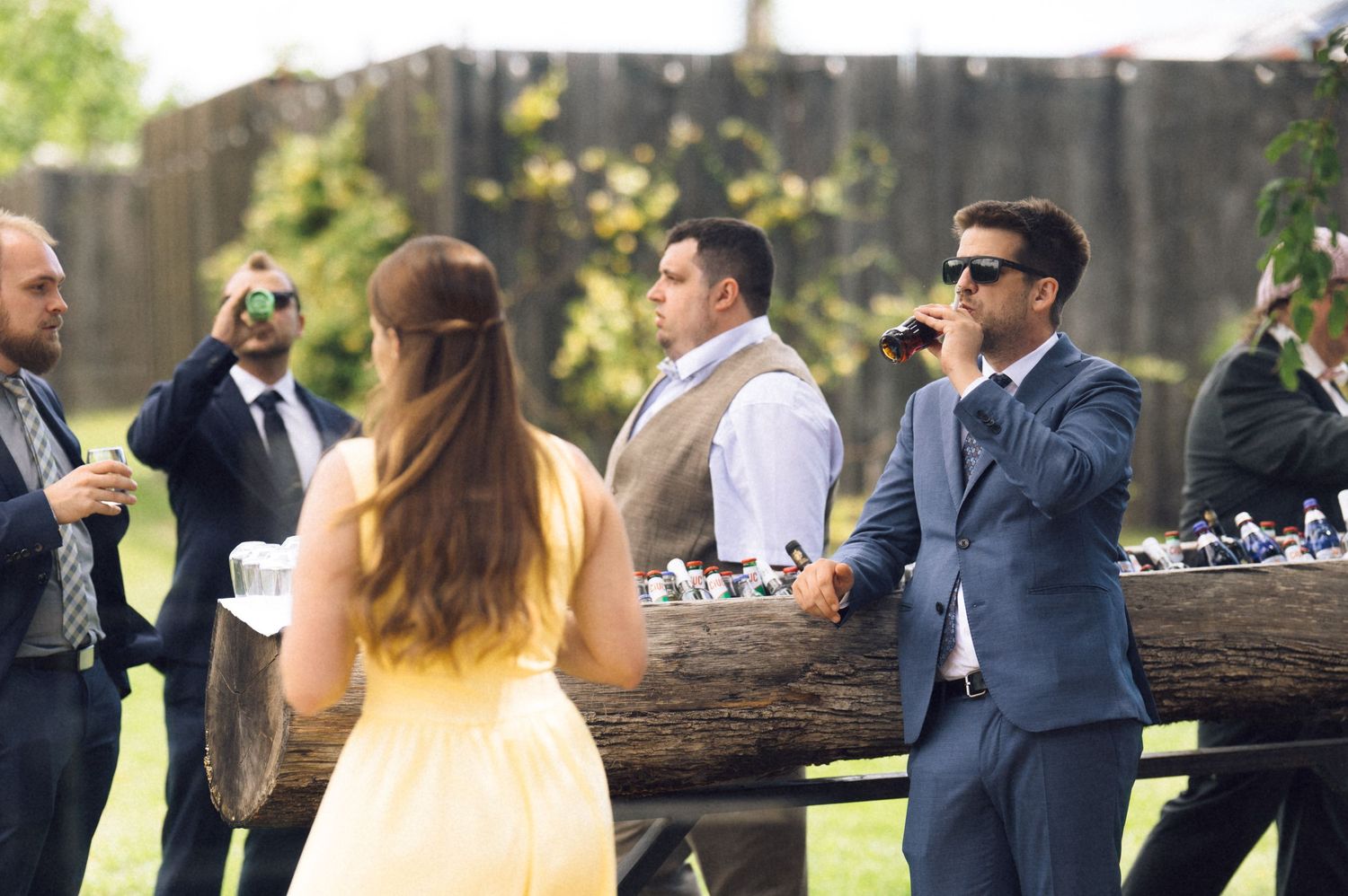 Wedding guests mingling outdoors, with men in suits and a woman in yellow dress at reception.
