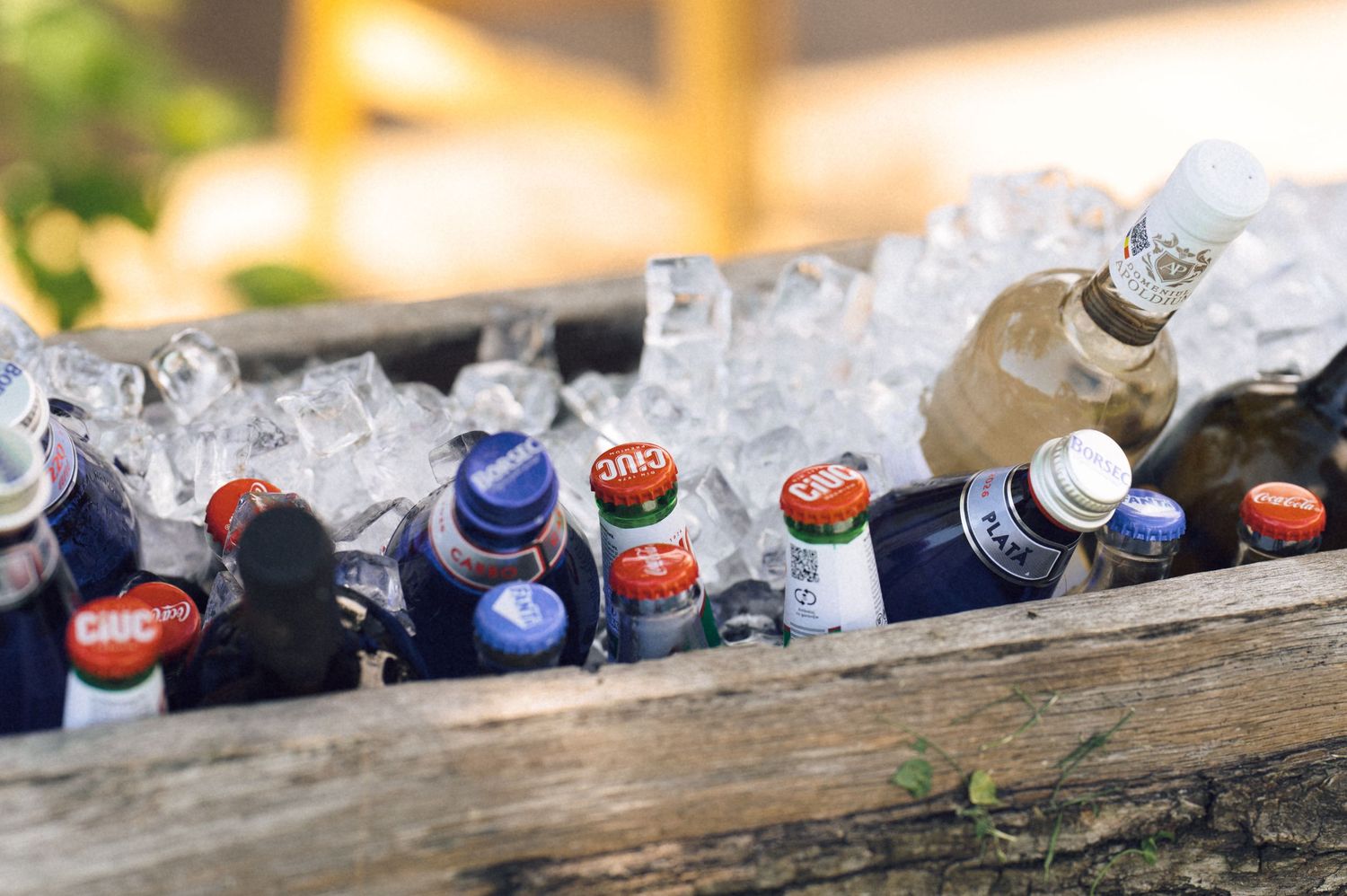 Assorted cold beverages and beer bottles chilling in ice inside a rustic wooden cooler box outdoors.