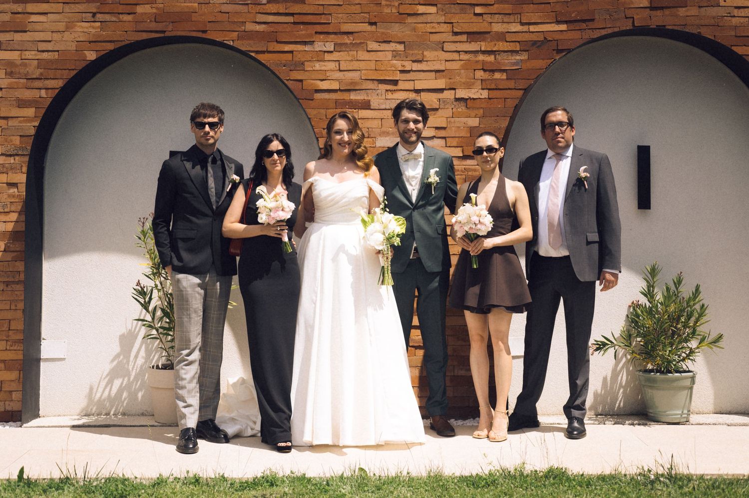 Wedding party of six posing in front of arched brick wall, bride in white gown with groom and guests.