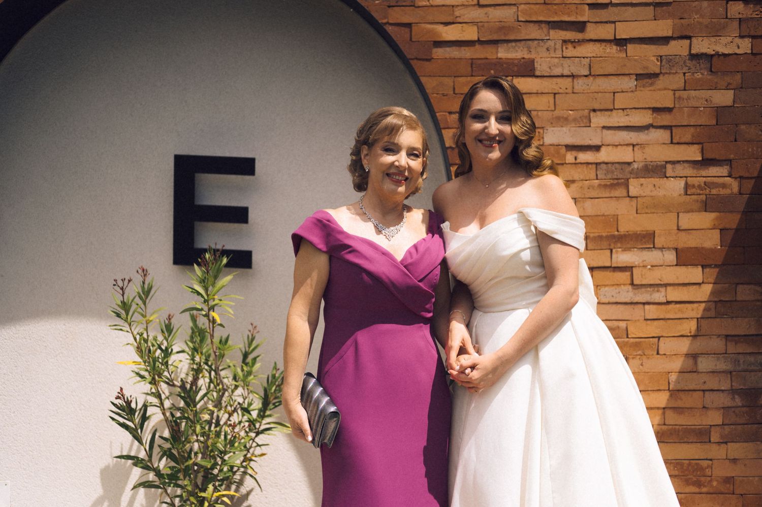Bride in white off-shoulder gown poses with woman in pink dress at elegant venue with brick wall.