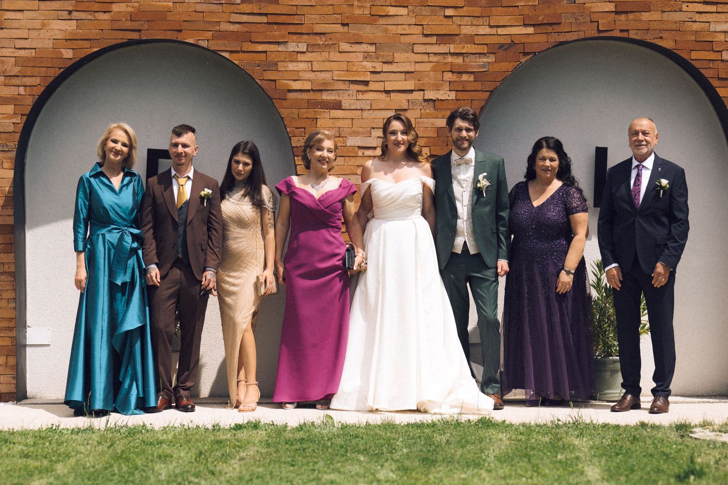 Wedding party of seven people posing outdoors in formal attire, including a bride in white gown, against brick arched wall.