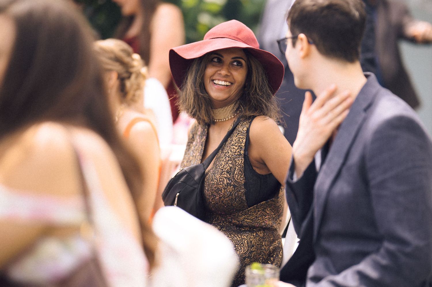 Woman in red wide-brim hat and lace dress smiling at a social gathering outdoors.