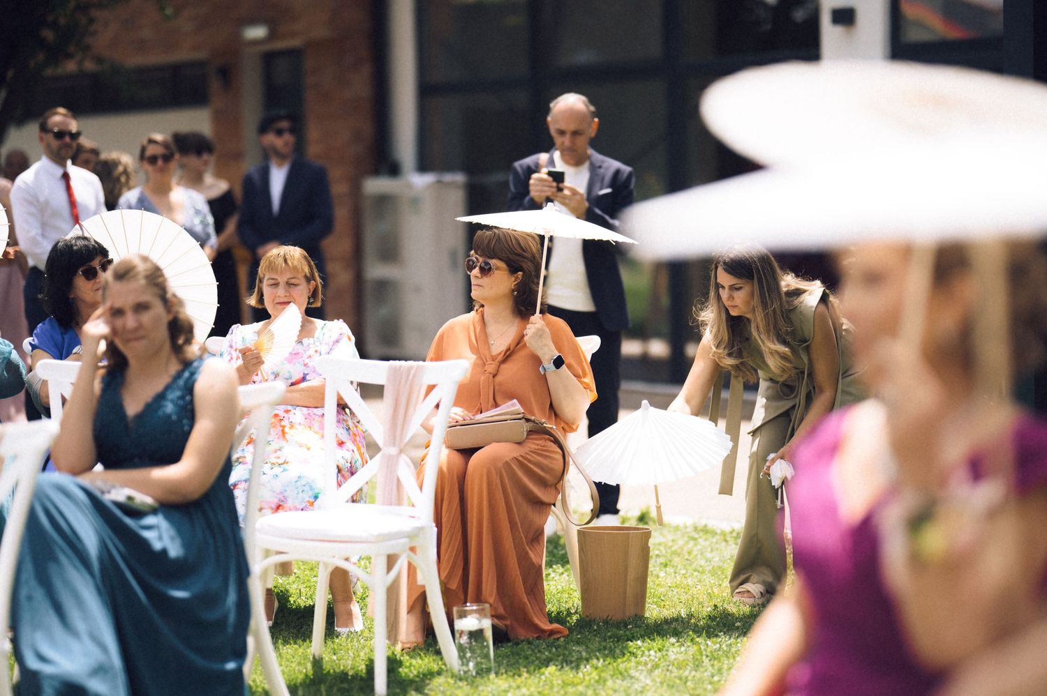 Guests in elegant attire seated on white chairs at an outdoor wedding ceremony on a sunny day.