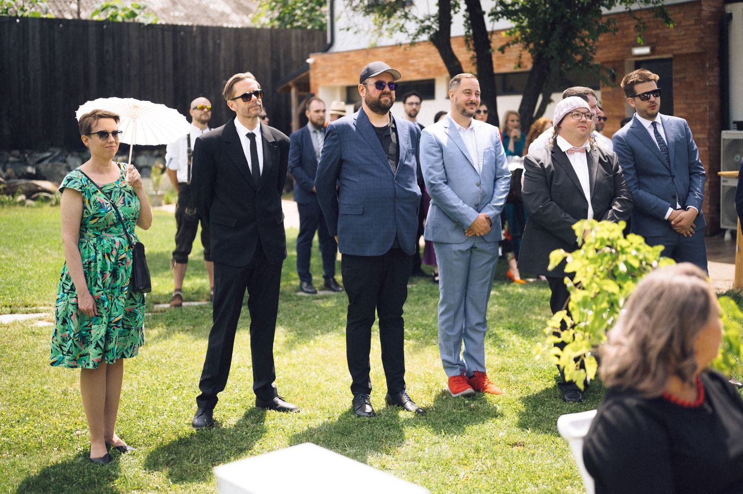 Group of elegantly dressed guests standing outdoors at a summer wedding ceremony on a sunny day.