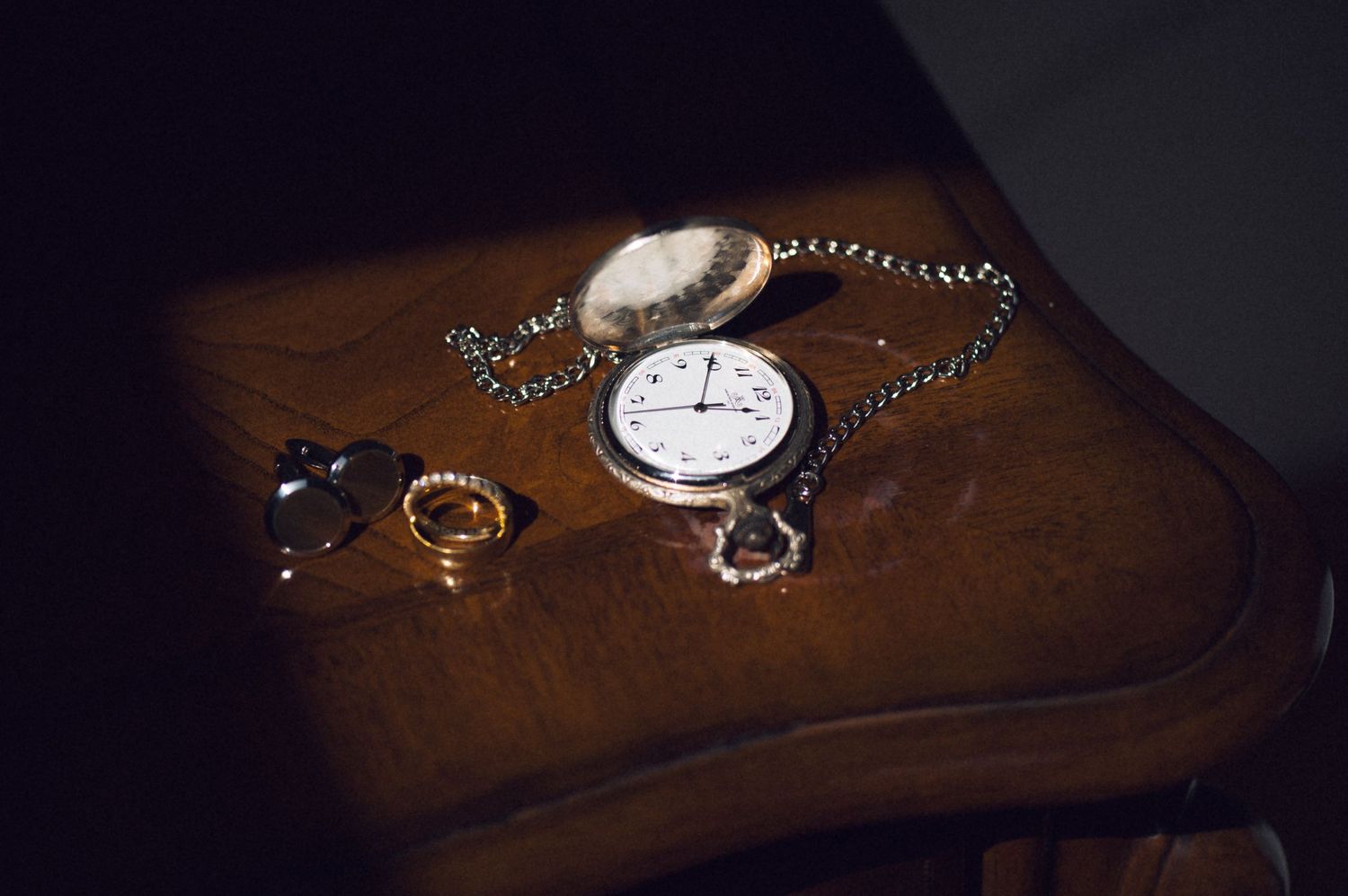 Vintage silver pocket watch with chain beside a gold ring and heart charm on a wooden surface.