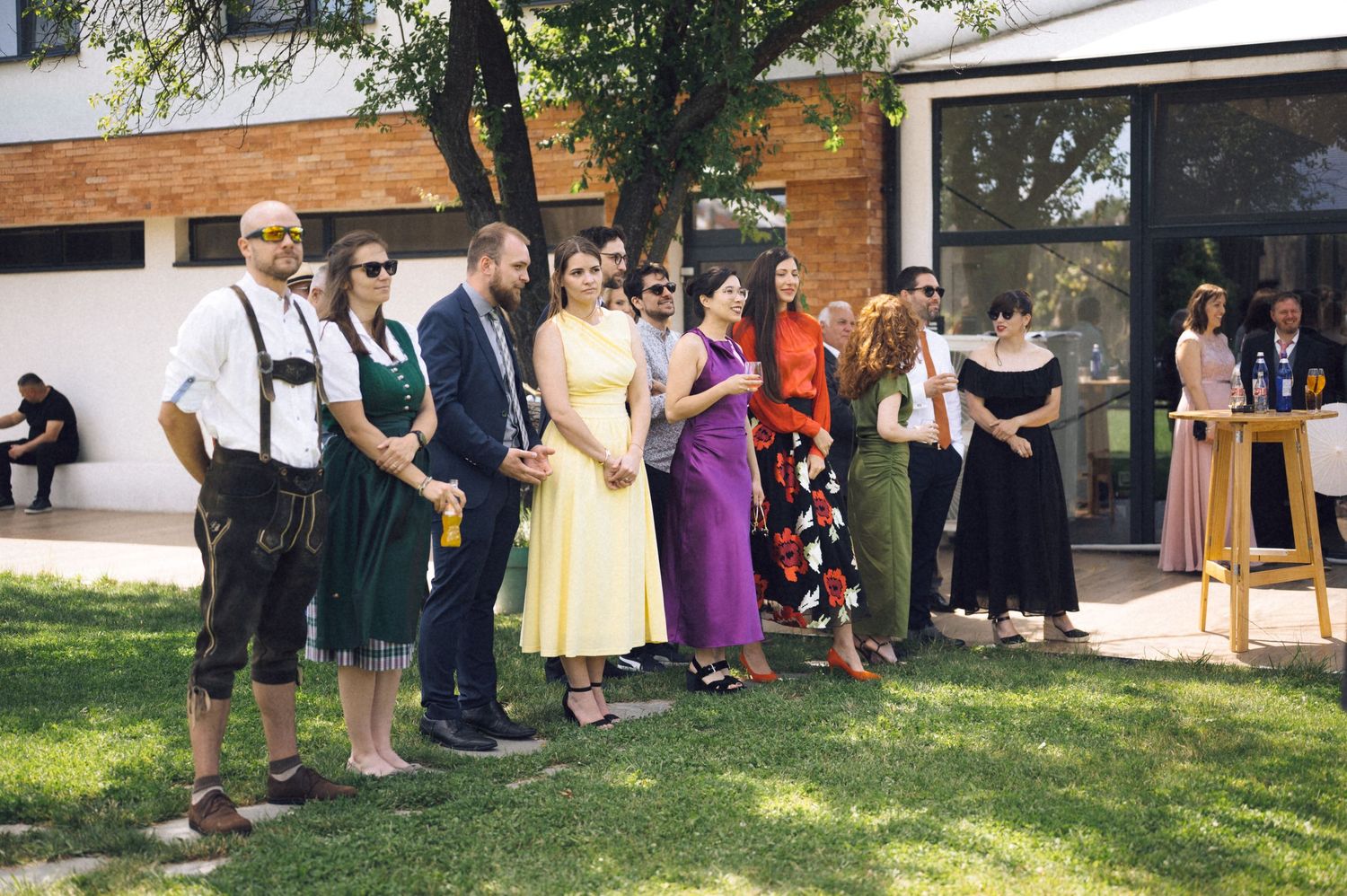 Group of people in colorful formal attire standing outdoors on green lawn near modern white building on sunny day.