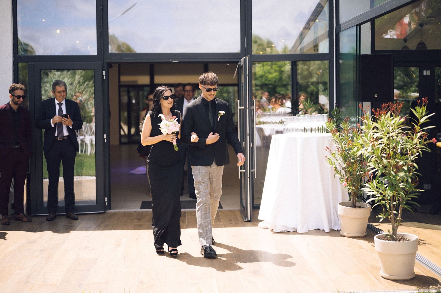 Couple in formal attire walking out of a modern venue entrance during a wedding celebration outdoors.