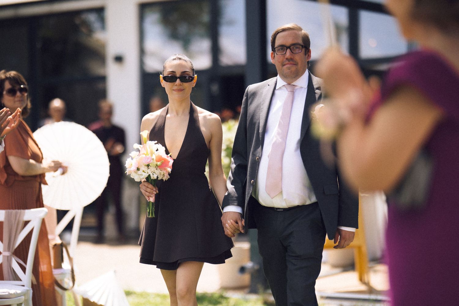 Couple walking hand in hand at outdoor wedding, woman in black dress holding pink bouquet, man in gray suit with pink tie.