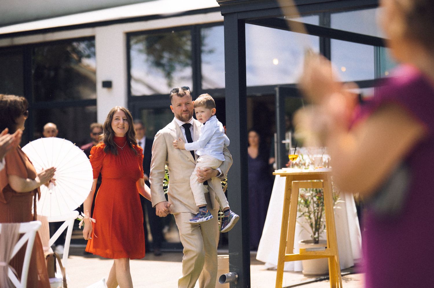 A man in a tan suit carries a young child while walking into an outdoor wedding venue with guests nearby.