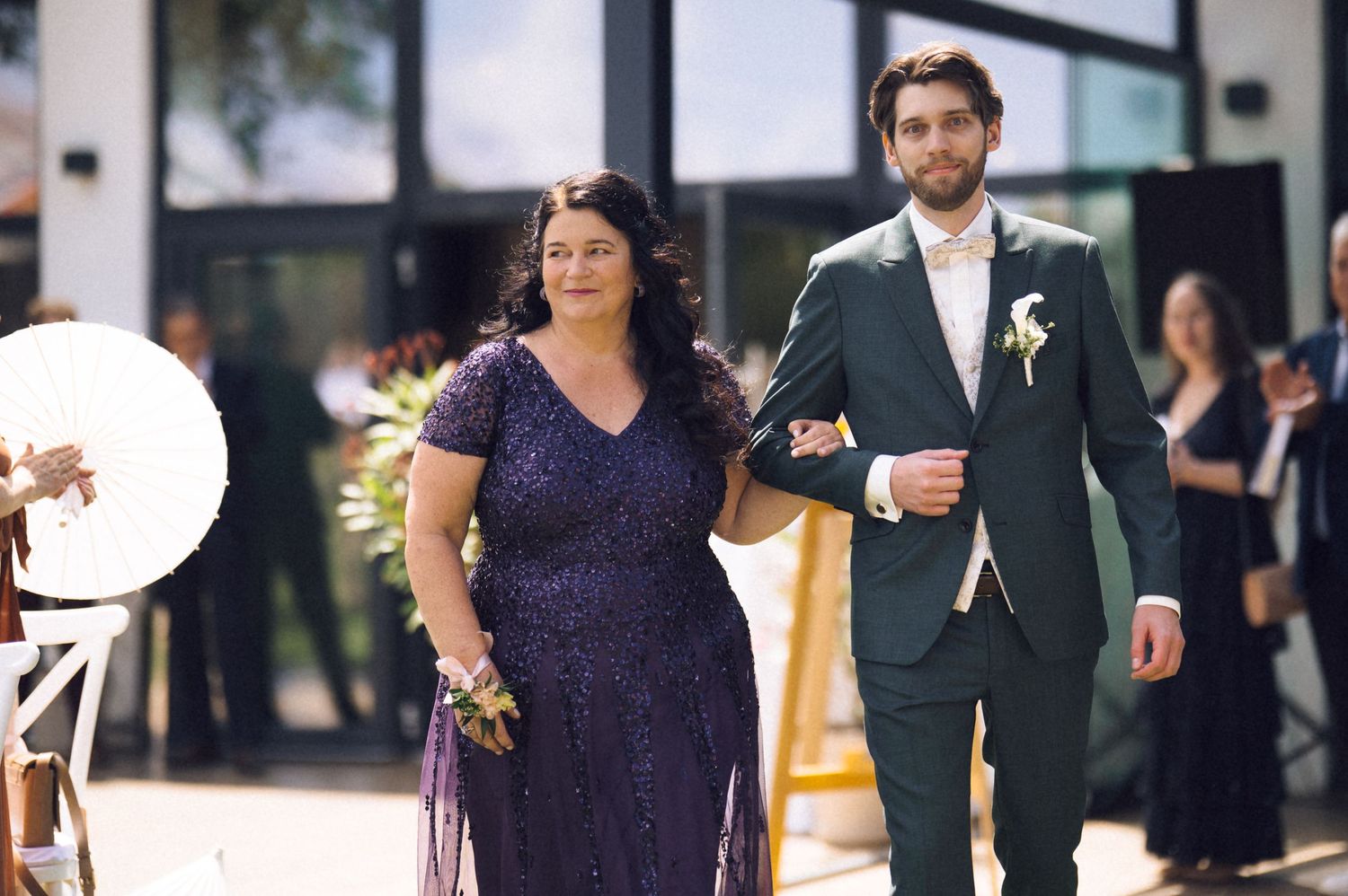 A woman in a purple sequin gown walks arm-in-arm with a man in a dark green suit at a wedding ceremony.