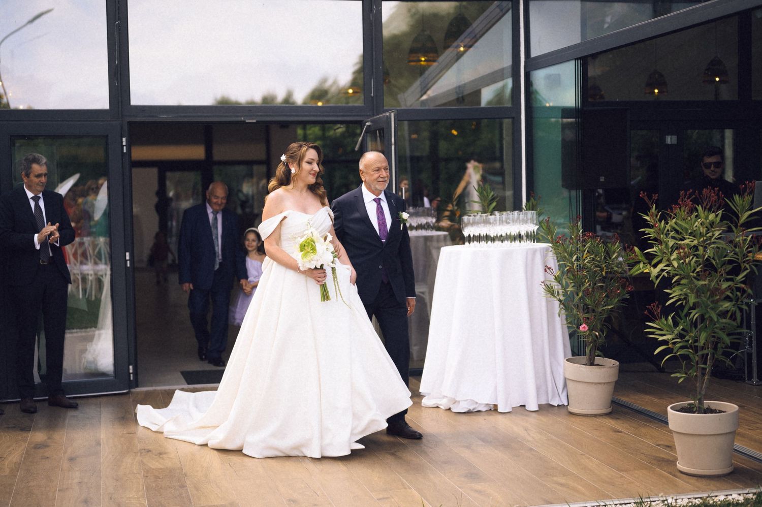 Bride in white gown walks with escort through elegant wedding venue entrance with guests and floral arrangements.