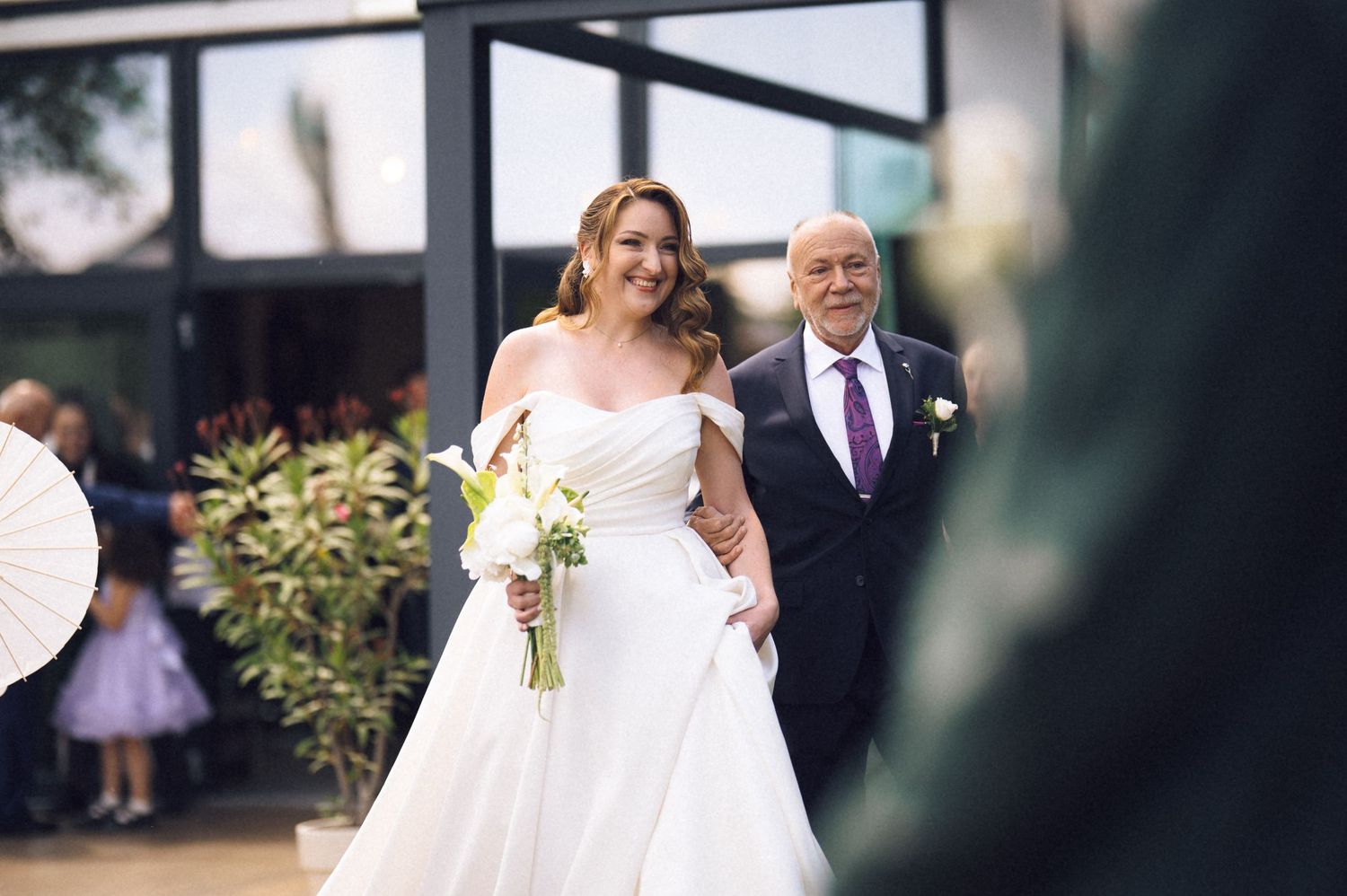 Bride in off-shoulder white gown walks down aisle holding bouquet with older gentleman in suit.