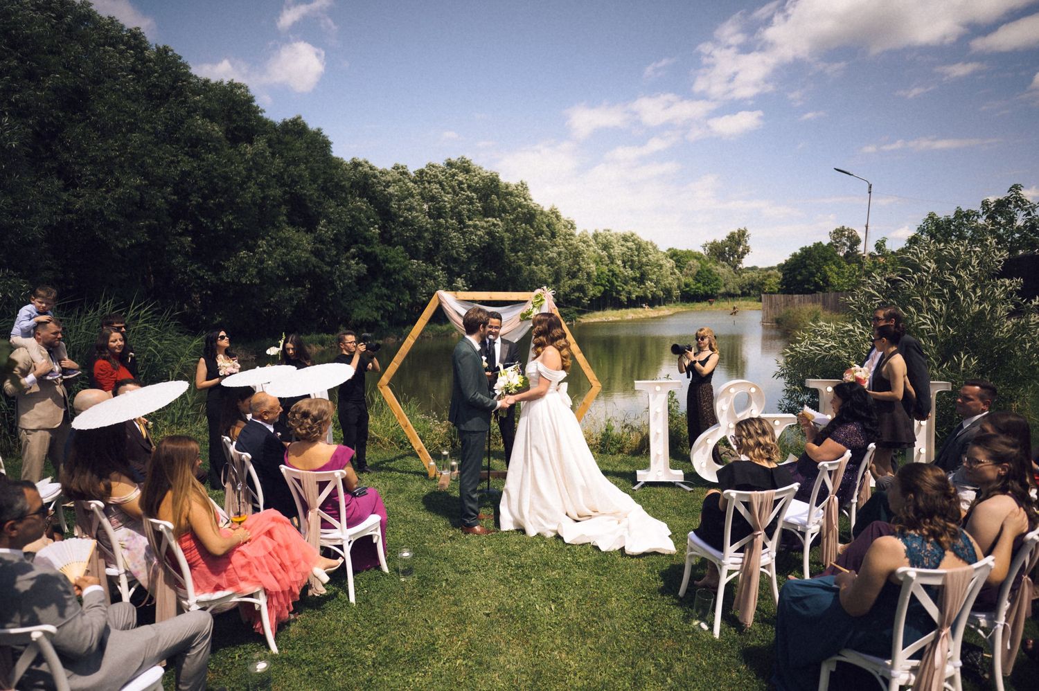 Outdoor wedding ceremony with bride and groom at geometric arch, guests seated on white chairs beside a scenic pond.