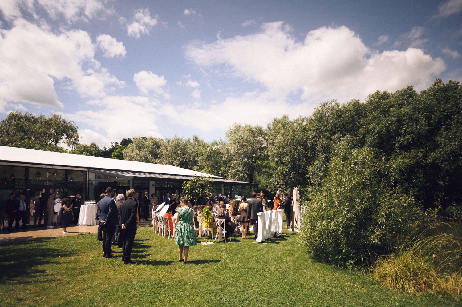 Guests mingle outdoors on green lawn beside a white marquee venue surrounded by lush trees under a partly cloudy sky.