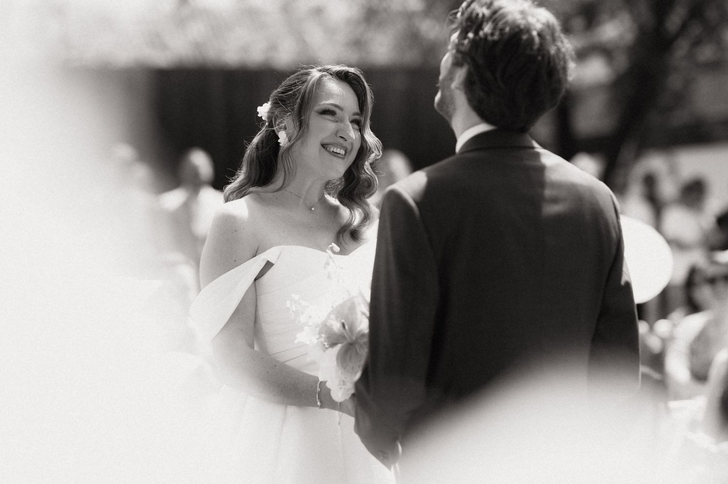 Black and white photo of smiling bride exchanging vows with groom at outdoor wedding ceremony.