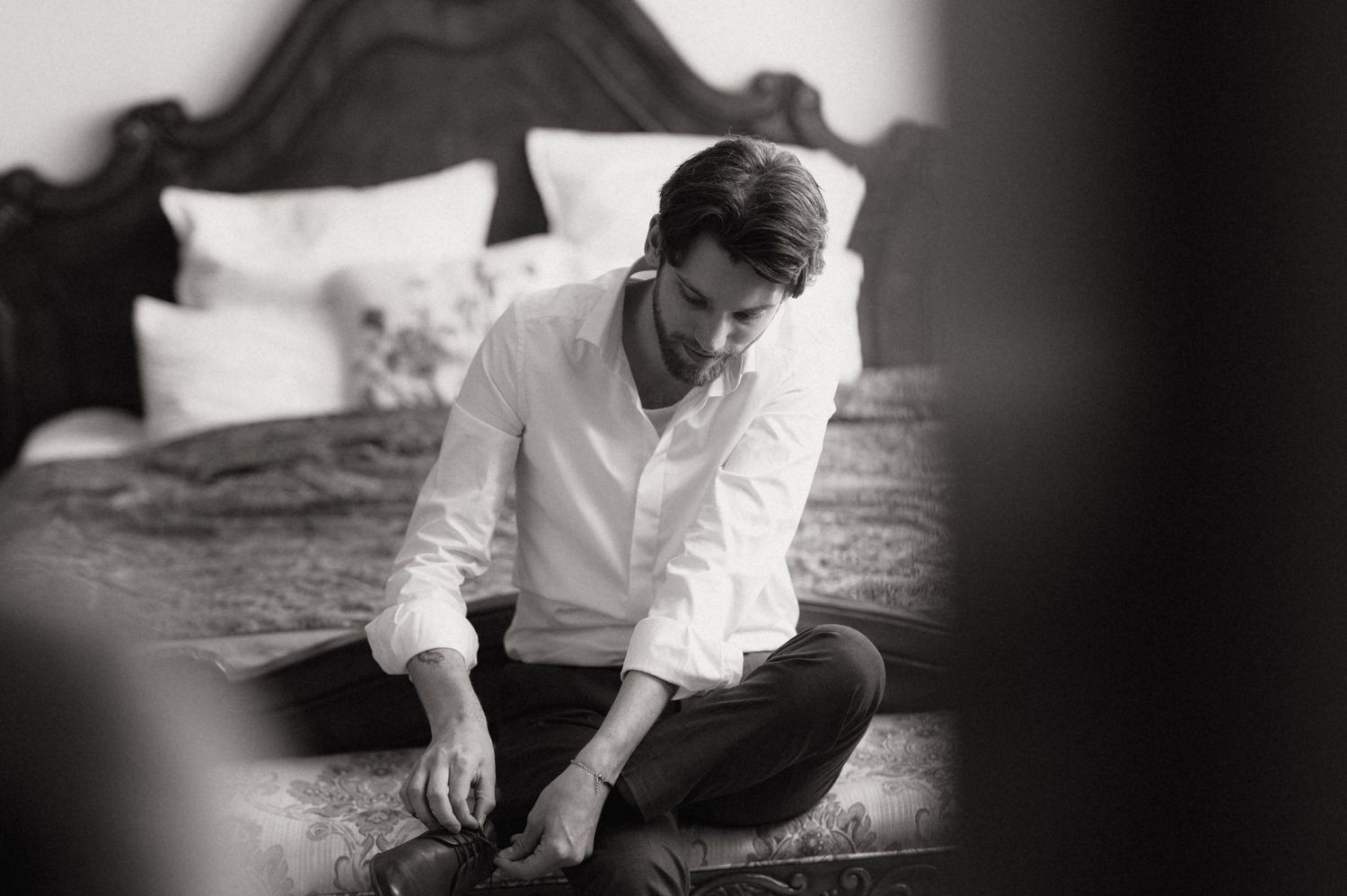 Black and white photo of a young man in white shirt sitting cross-legged on a bed, looking down thoughtfully.