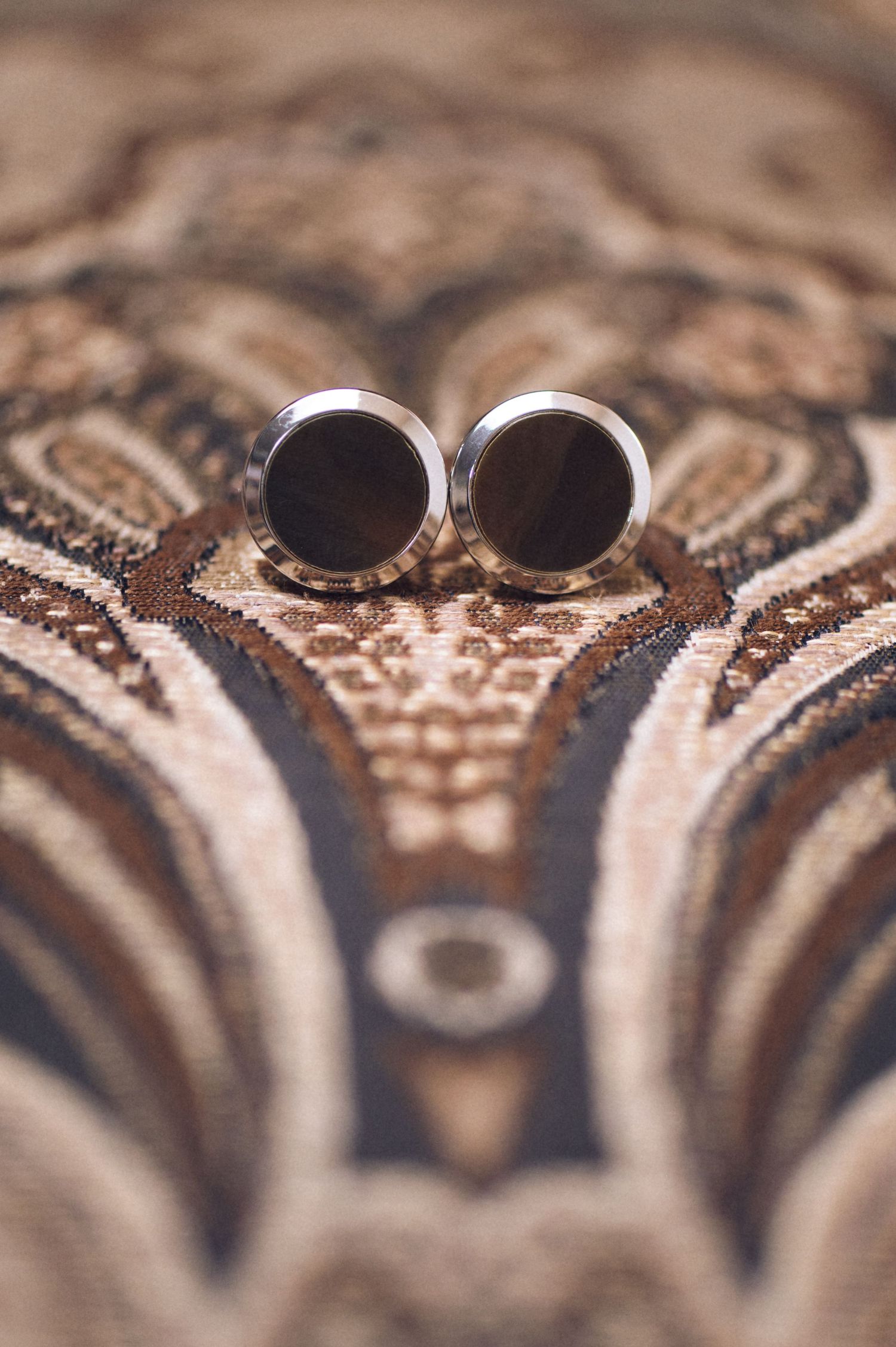 Elegant round dark wood cufflinks with silver trim displayed on ornate patterned fabric.