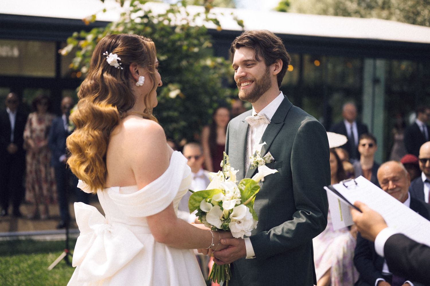 Bride holding white bouquet smiles at groom in dark suit during outdoor wedding ceremony.