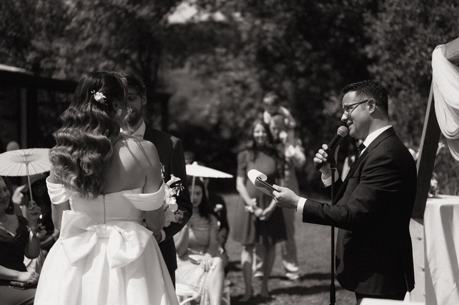 Outdoor wedding ceremony in black and white, groom reads vows to bride while guests watch in background.