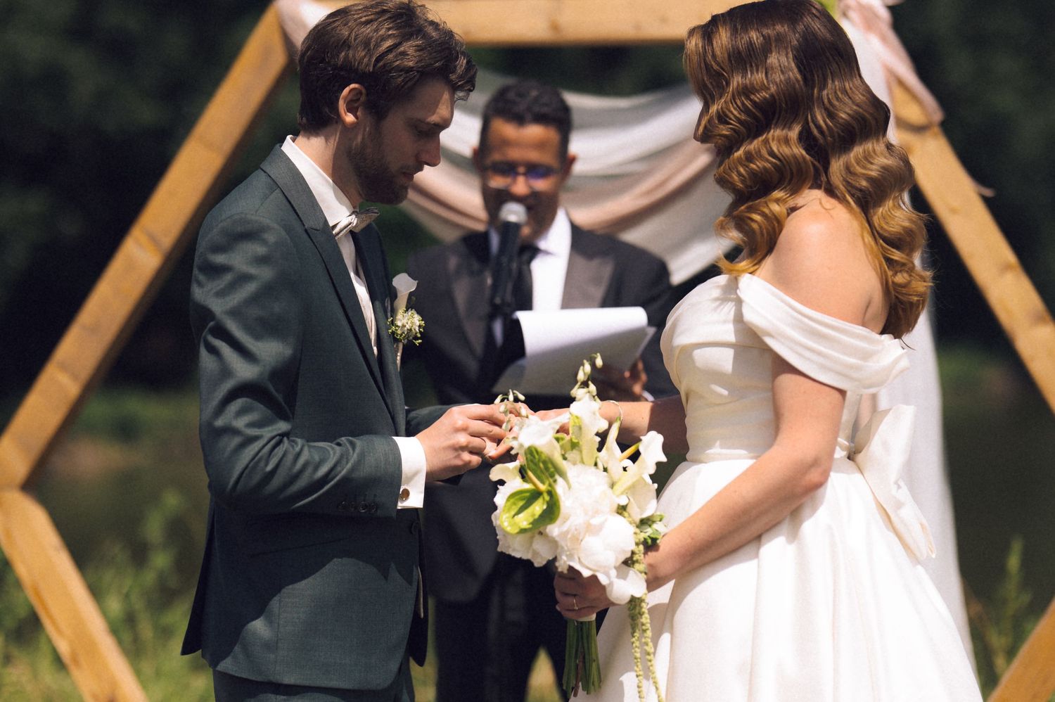 Groom placing wedding ring on bride's finger during outdoor ceremony with officiant and hexagonal wooden arch.