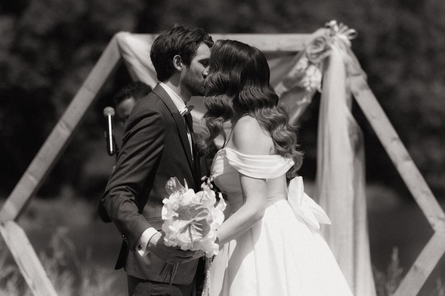 Black and white photo of a bride and groom sharing their first kiss at an outdoor wedding ceremony.