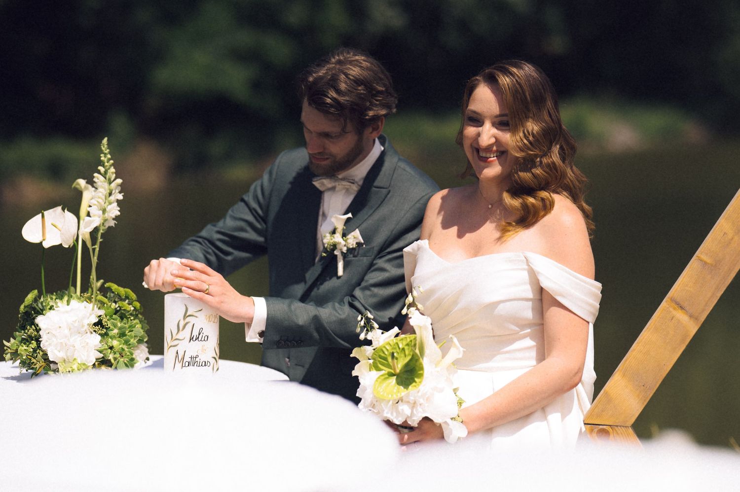 Bride and groom smiling at outdoor wedding ceremony table with white flowers near a serene lake backdrop.