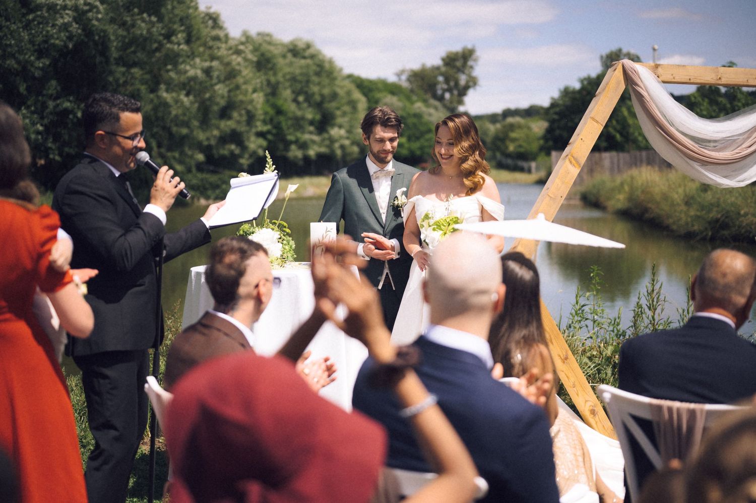 Outdoor wedding ceremony by a river with couple exchanging rings, officiant speaking, and guests watching in sunny weather.