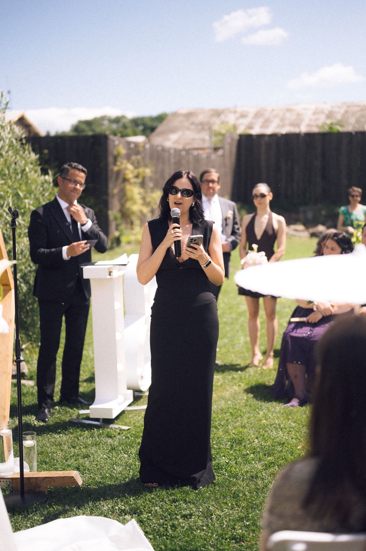 Woman in black dress speaks at outdoor wedding ceremony, holding microphone and phone, guests seated on sunny lawn.