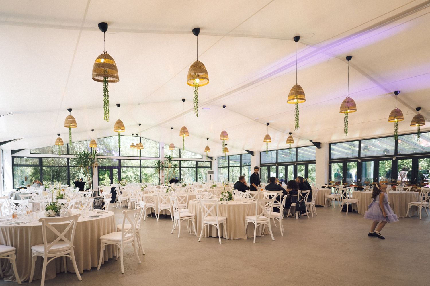 Elegant wedding reception venue with round tables, white chairs, hanging gold pendant lights, and large windows overlooking greenery.