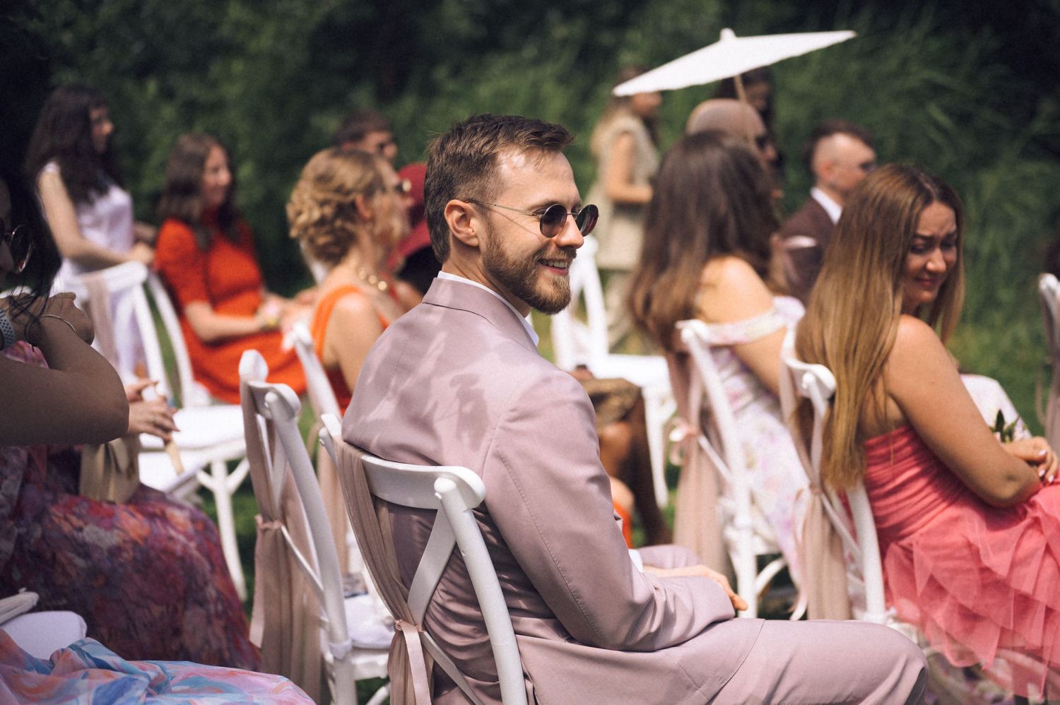 Man in sunglasses smiling at outdoor wedding ceremony seated among guests in summer garden setting.