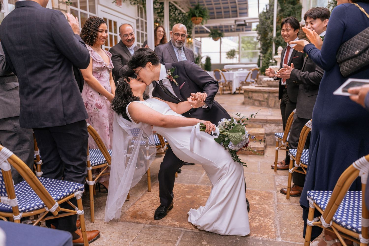 Groom carries bride in wedding dress on urban street with red bus and brick hotel building in background.