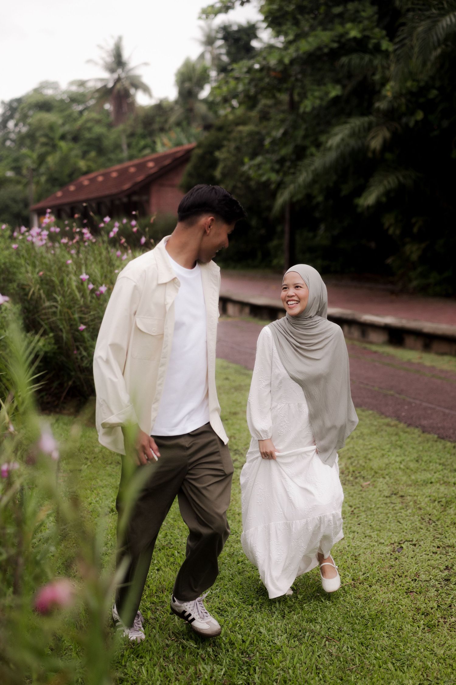 Muslim couple walking hand in hand in a lush garden, woman in hijab and white dress, man in cream jacket.