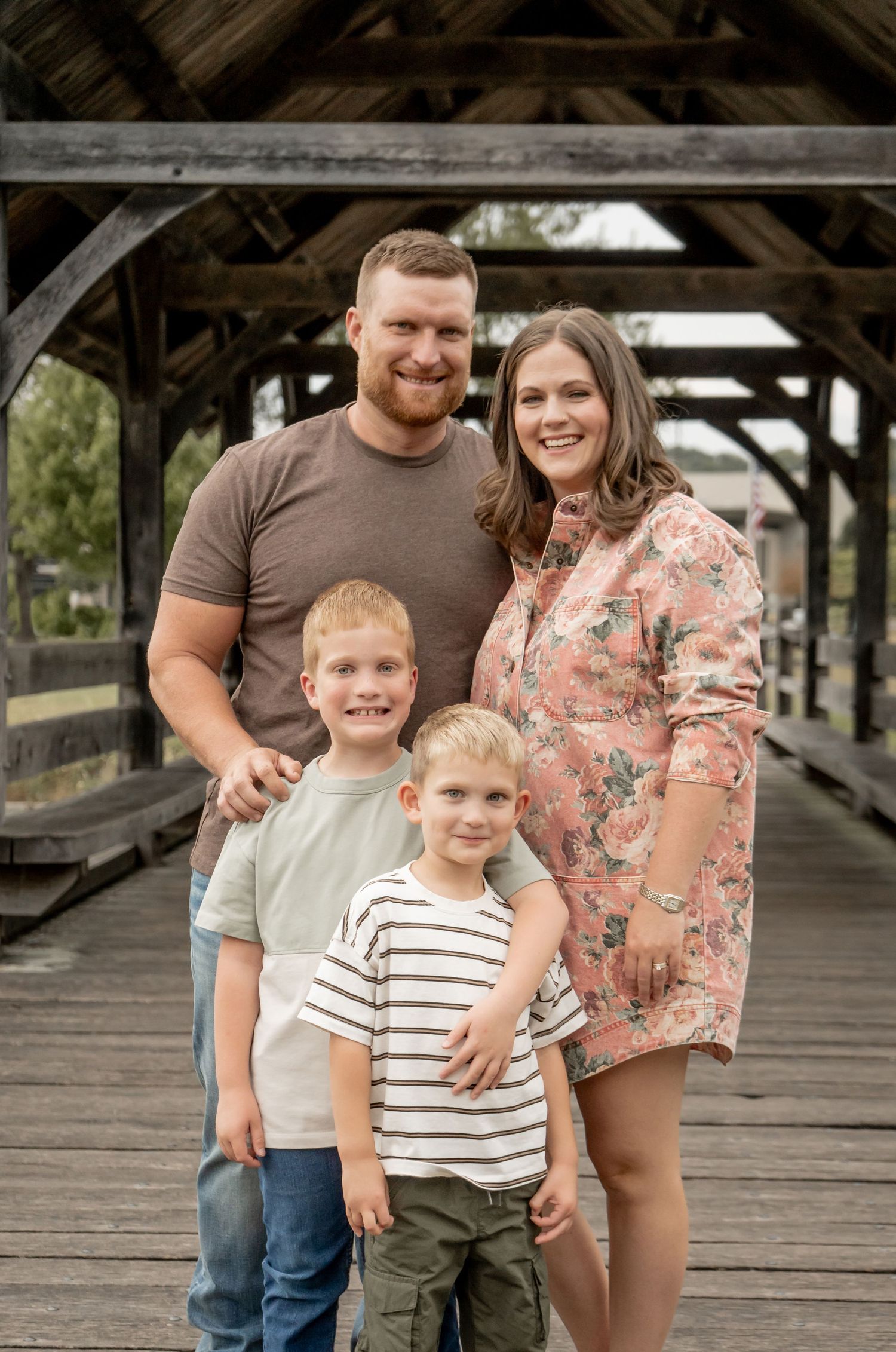 Smiling family of four posing together on a rustic wooden covered bridge outdoors.