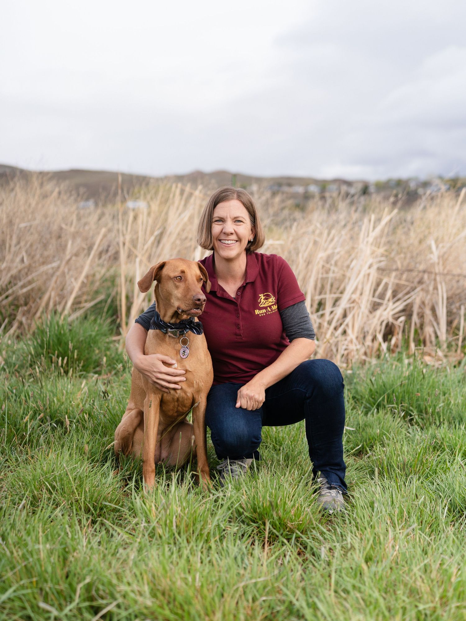 Woman in maroon polo shirt kneeling beside a brown dog on green grass with tall reeds in the background.