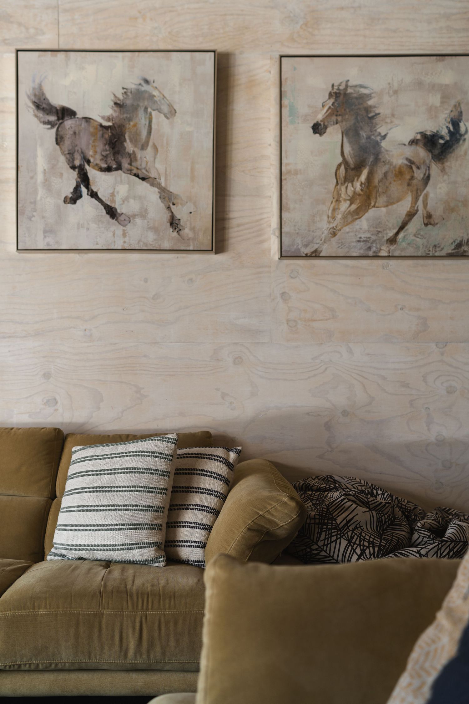 Olive velvet sofa with striped pillows beneath two framed equestrian horse paintings on a light plywood feature wall.
Cedar & Stone