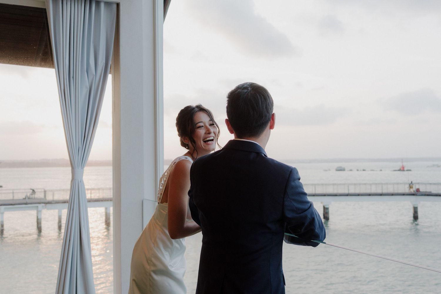 Bride laughing joyfully with groom during first look at waterfront wedding venue with ocean views.