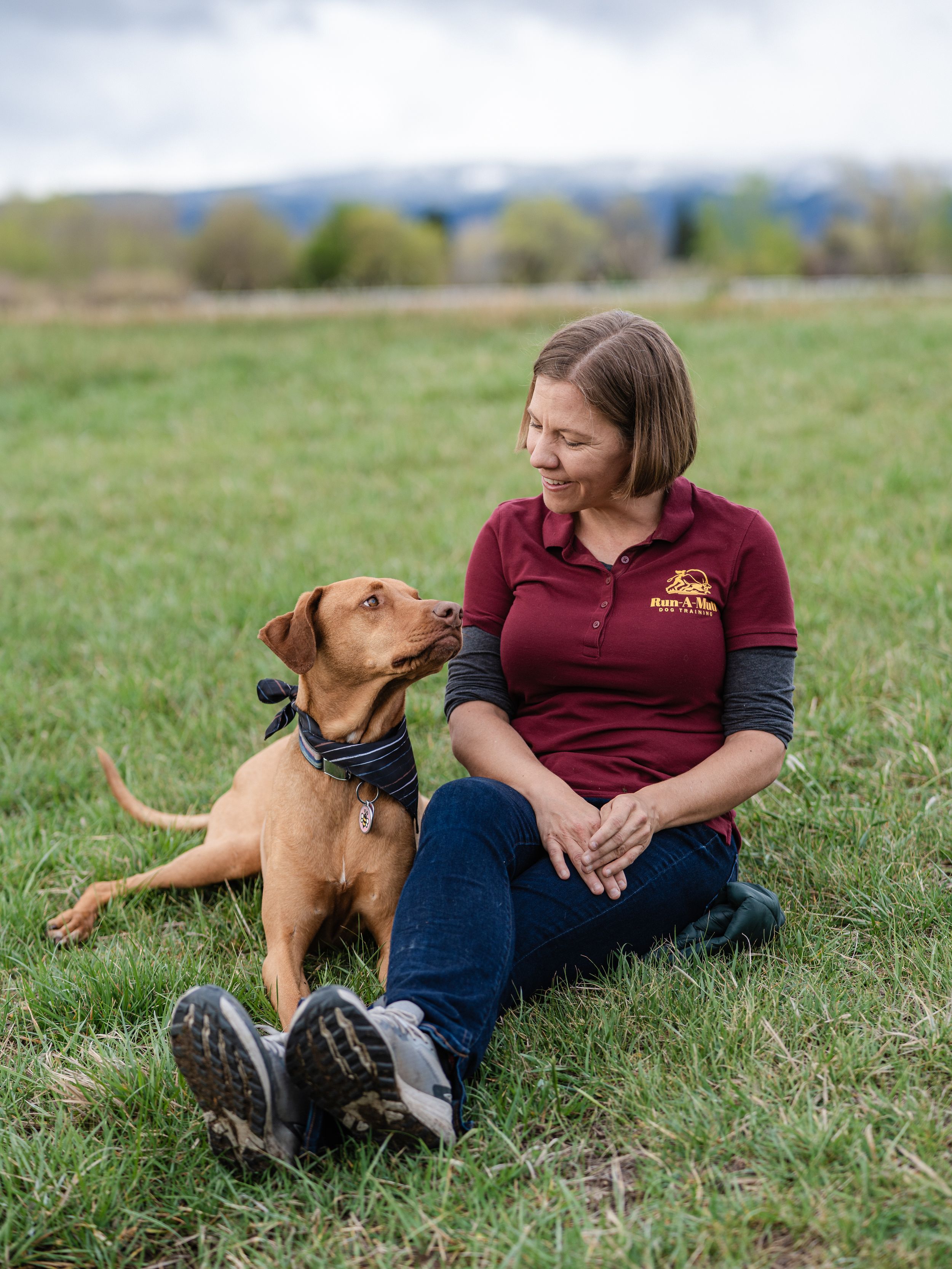 Woman in maroon polo sits on grass with a brown dog looking up at her in an open field.