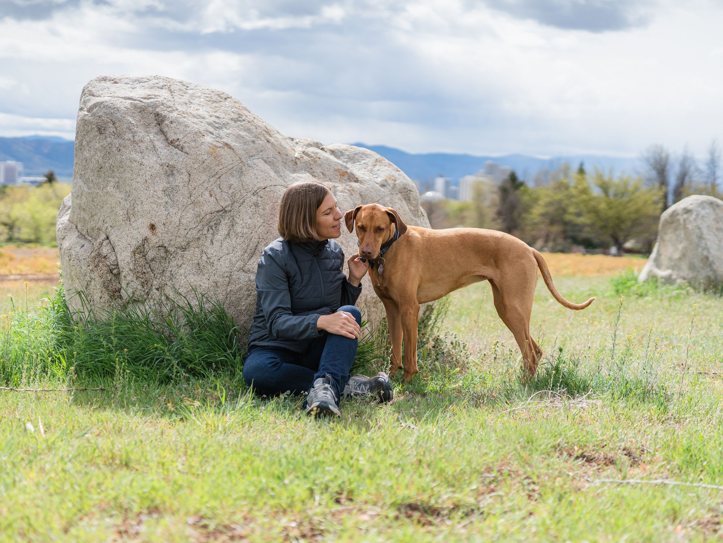 Woman sitting on grass beside a large rock, sharing a tender moment with her Rhodesian Ridgeback dog outdoors.
