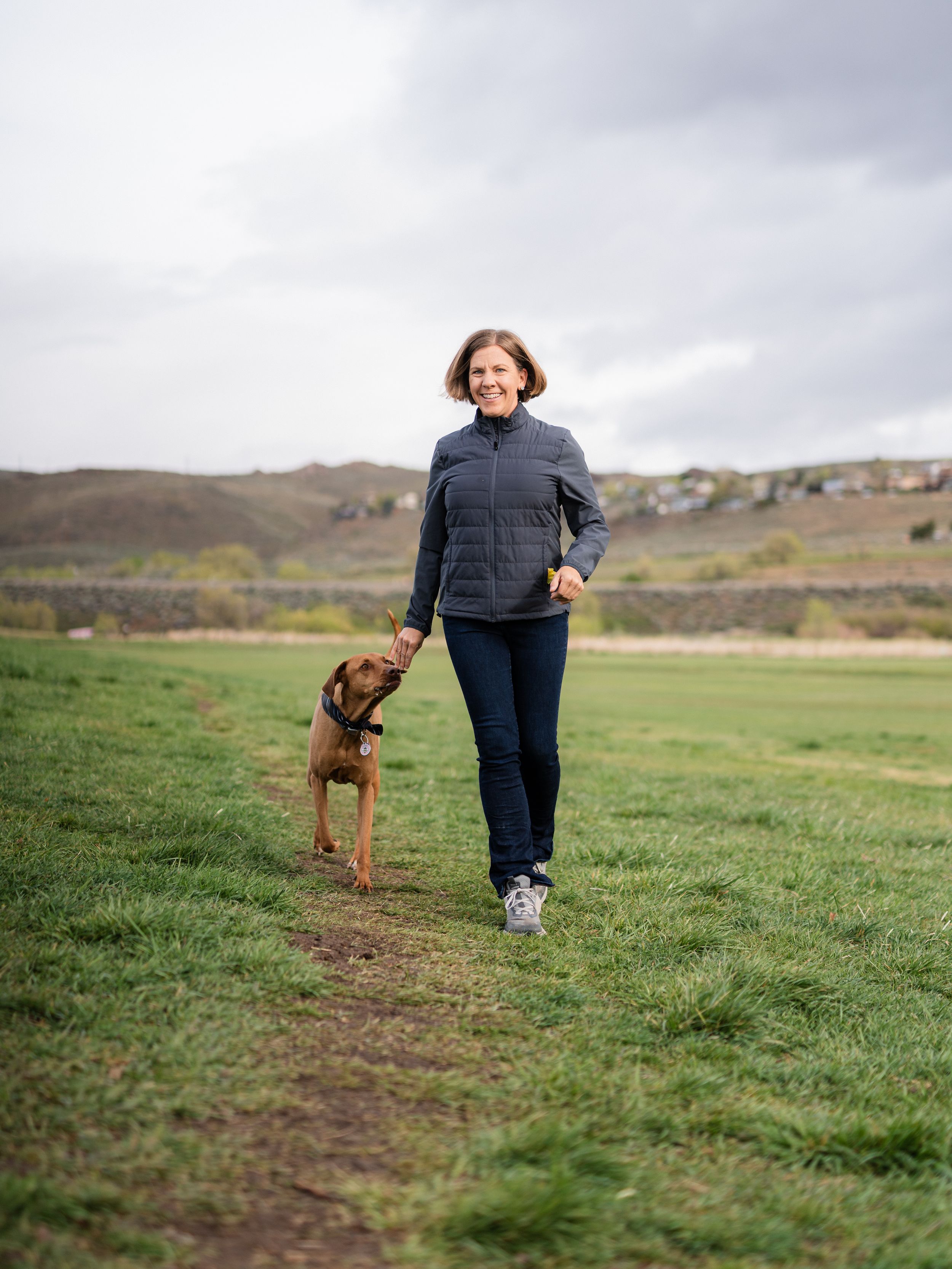 Woman walking a brown dog on a grassy path in an open field with rolling hills in the background.