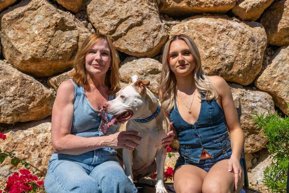 Two women sitting outdoors with a white dog in front of a stone wall, surrounded by red flowers on a sunny day.