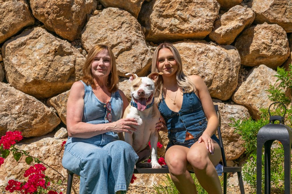 Two women in denim outfits sit on a bench with a white dog in front of a stone wall with red flowers.