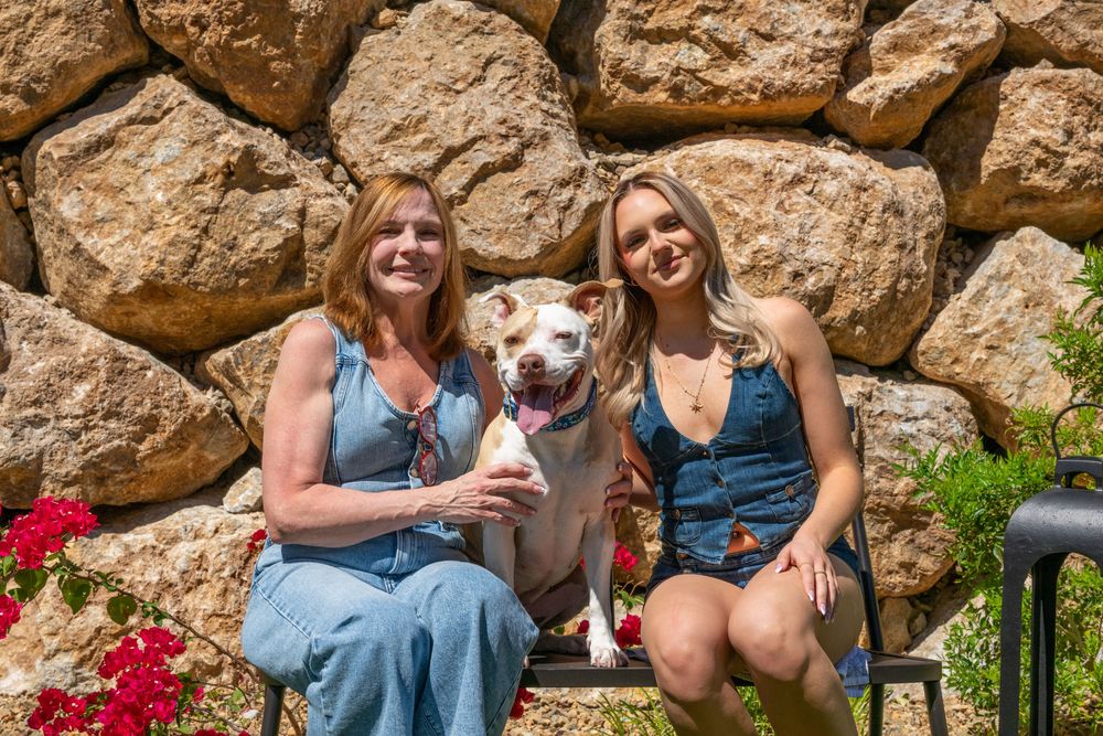 Two women and a pit bull dog pose together in front of a stone wall with red flowers.